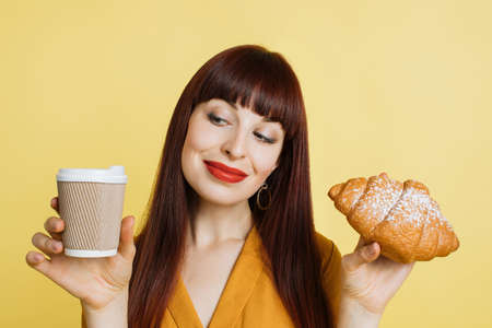 Closeup portrait of glamorous attractive young woman with bright makeup, posing on yellow studio background and holding take away coffee cup and fresh tasty croissant. French breakfast.の写真素材