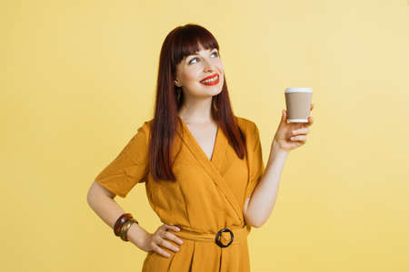 Enjoying the coffee break. Pretty dreamy smiling woman in bright yellow overalls with take away coffee cup, posing on isolated yellow studio background and looking up, copy space.の写真素材