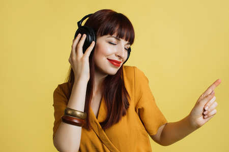 Charming gorgeous brunette girl in yellow clothes and earphones, listening to music and dancing with eyes closed posing on yellow background. People, music, emotions concept.の写真素材