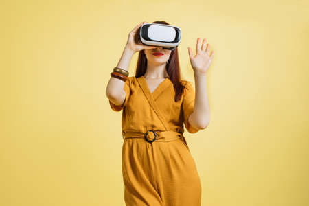 Excited young Caucasian woman in VR headset touching air exploring virtual reality at studio yellow background. Pretty lady enjoying artificial computer world or playing video game.の写真素材