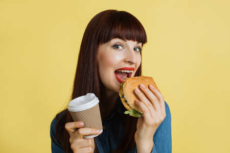 Portrait of young red haired hungry woman holding take away coffee cup and biting fresh hamburger, smiling to camera over isolated yellow background. Lunch, diet and jank food conceptの写真素材