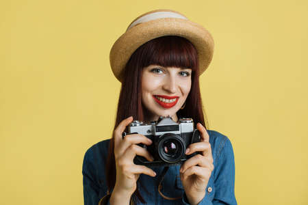 Stylish red haired smiling woman photographer , wearing straw hat and denim shirt, posing with retro camera on the yellow wall background. Image with copy space.の写真素材