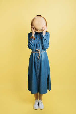 Portrait of attractive young trendy travel woman, wearing jean dress, hiding face with stylish summer straw hat, posing on camera in studio isolated over yellow backgroundの写真素材