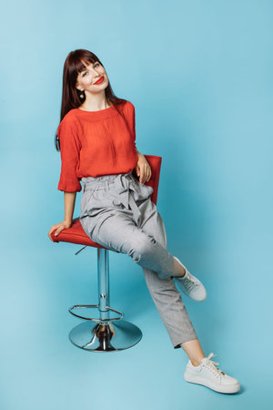 Pretty smiling Caucasian woman in stylish clothes, sitting on chair against blue background. Full length portrait of young businesslady with bright makeup, sitting on the chair.の写真素材