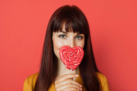 Close up studio shot of charming young red haired woman, wearing yellow shirt, holding a heart shaped red sweet lollipop, covering her nose and lips, posing on a red background.の写真素材