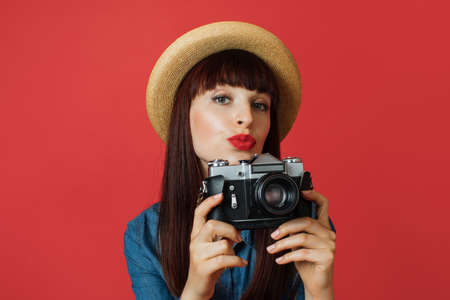 Hobby, taking photos, travel concept. Gorgeous young woman in hat, photographer amateur, smiling and making air kiss, taking picture on old vintage camera, on colorful red studio backgroundの写真素材