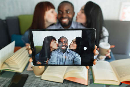 Three multiethnic friends, enjoying time for reading books in cafe together, taking selfie photo on dogotal tablet. African man grinning to camera while Caucasian and Asian girl kissing him on cheeksの写真素材