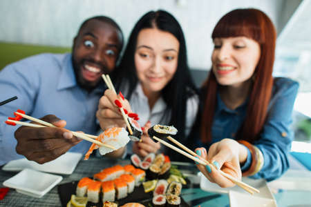 Blurred view of smiling pleasant diverse mixed race friends, having fun and enjoying lunch in Chinese restaurant, holding wooden sticks with tasty delicious sushi rolls. Focus on hands with rollsの写真素材
