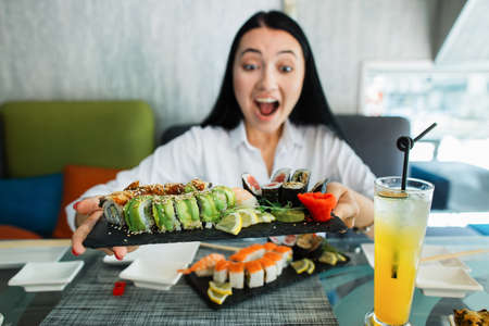 Wow, its tasty. Close up of excited brunette asian woman happy to have delicious lunch in asian restaurant and eat sushi rolls. Focus on big black plate with colorful rolls in hands of womanの写真素材