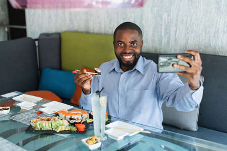 Excited smiling young African business man doing selfie shot or video call on mobile phone, holding chopsticks with sushi roll, having traditional oriental lunch at restaurantの写真素材