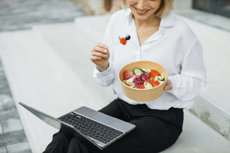Close up view of hand of businesswomann having a vegetables salad for lunch, healthy eating sitting on stairs using modern laptop, lifestyle concept, unrecognizable person.の写真素材