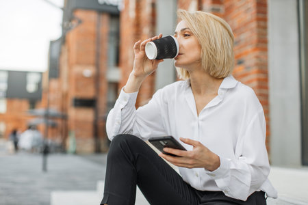 Cheerful young blond woman wearing white shirt and black pants sitting on stairs outdoors, drinking takeaway coffee cup, using mobile phone.の写真素材