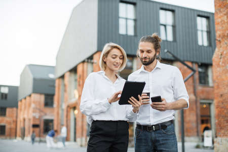 Charming woman with digital tablet and attractive man with smartphone and cuo of take away coffee standing together near office building. Two colleagues using modern gadgets for work outdoors.の写真素材
