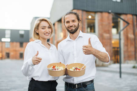 Young cheerful couple, colleague or business partners in white shirts having break and eating healthy meal walking at the city street showing thumb up. Organic healthy nutrition concept.の写真素材