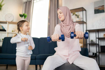 Focus on dumbbells, young Arab family doing sports in a bright living room at the weekend. Happy free time, healthy lifestyle concept.の写真素材