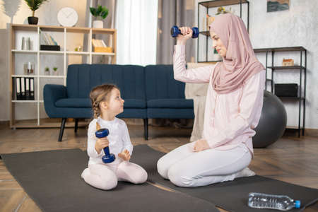 Cute little girl doing sports with her Muslim mother in a hijab sitting on a mat and doing exercises with dumbbells in living room at home.の写真素材