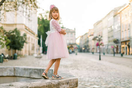 Fashion full length portrait of a cute little blond girl, wearing pink dress and flower in hair, posing to camera, while standing on ancient stone fountain in old European city.の写真素材
