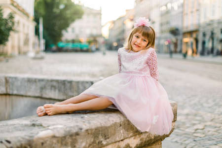 Romantic portrait of charming Caucasian little girl in trendy pink princess dress and without shoes, sitting on the old stone fountain on the street on ancient city background.の写真素材