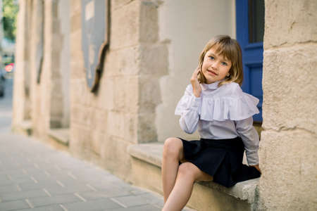 Outdoor fashion street city portrait of little Caucasian girl. Happy little girl sitting near the old building wall in ancient European city with blue door on the background.の写真素材