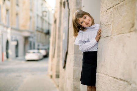 Cute little Caucasian blond girl in black and white clothes, posing to camera outdoors in the old city street, hiding her face behind the ancient building wall.の写真素材