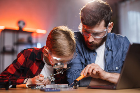 Little boy in eyeglasses soldering display card while caring father sitting near and helping him....