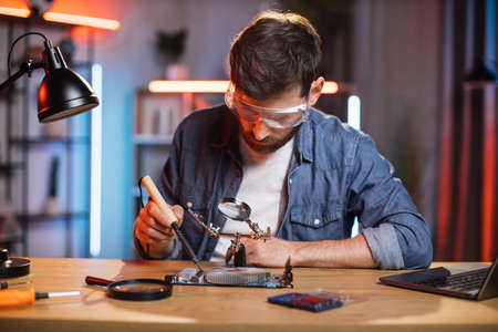 Focused bearded man in protective glasses sitting at table and soldering video card from modern laptop. Young male repairing important detail from computer by himself at home.の写真素材