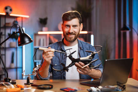 Smiling happy Caucasian bearded guy using soldering iron for repair modern quadcopter at home. Young man in casual clothes sitting at table and repairing drone.の写真素材