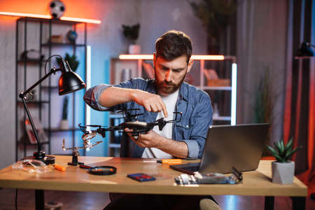 Concentrated bearded man using various tools for repairing modern quadcopter at home. Young caucasian guy in denim shirt fixing flying drone by himself.の写真素材
