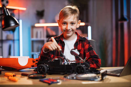 Portrait of smiling little boy sitting at table and soldering remote controlled red car. Caucasian child in casual wear fixing favorite toy by himself.の写真素材