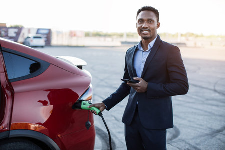Young successful businessman charging electric car on station and using modern smartphone for work....