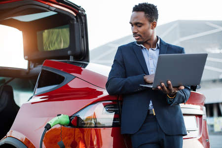 Successful african businessman in formal wear recharging battery of electric car and working on portable laptop.Young male standing outdoors near luxury red car with modern computer in handsの写真素材