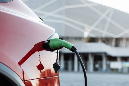 Close up of red luxury electric car charging at modern station. Green cable plugged into auto...