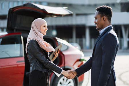 Competent female auto dealer holding digital tablet and shaking hands with male customer. Muslim woman in hijab selling luxury electric car to african businessman in suit.の写真素材