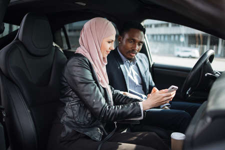 African american businessman in suit and muslim woman in hijab using modern smartphone for remote work while sitting together inside luxury car. Concept of people, gadgets and vehicle.の写真素材