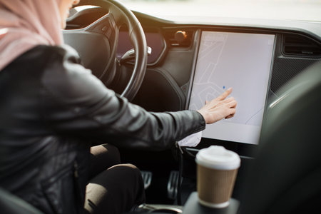 View from shoulder to arabian woman in hijab sitting on drivers seat of luxury electric car and...