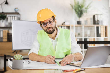 Architect man or male builder hold pen and ruler, writing and drawing architectural project, work on laptop computer on the desk at workplace looking at camera.の写真素材