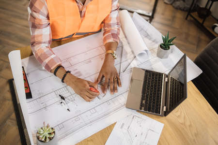 View from the top of table with tools for drawing. African designer in orange reflective vest draws the plan of the house. Hands of engineer. Architect sketching a construction project.の写真素材