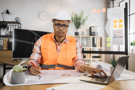 Portrait of african american man wearing uniform and white hard hat at work. Confident contractor sitting at the desk, holding pen and working on laptop with sketches of building object.の写真素材
