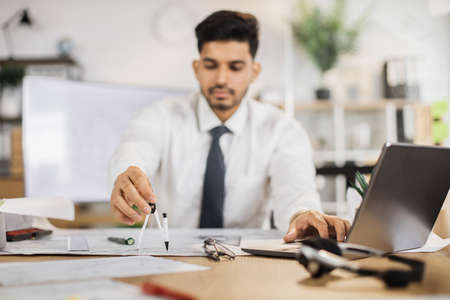 Focus on hand with compass. Young indian builder, engineer in white shirt with tie sitting on desk in office on background of large TV screen with , using laptop and looking on compass.の写真素材