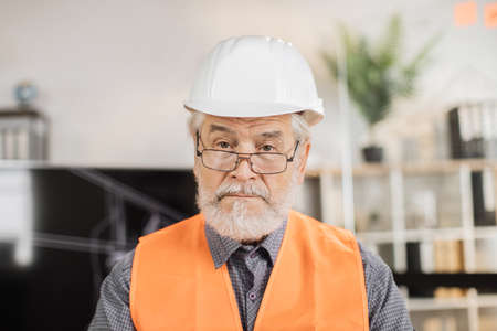 Portrait of mature architect wearing white construction helmet indoors. Bearded man in orange reflective vest posing among architectural office of the company.の写真素材