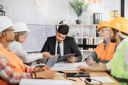 Indian businessman signs contract and documents of senior woman boss of a construction company. Diverse group of architects, designers and engineers sitting at table with modern gadgets and blueprintsの写真素材