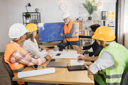 Mature builder caucasian man talks about the project on big lsd monitor screen with blueprints to his colleagues architects, builders and designers. Teamwork and brainstorming concept.の写真素材