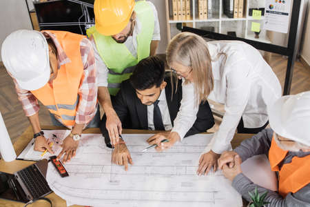 Top view of five diverse people in suits and helmets sitting at office with blueprints and gadgets. Qualified construction team planning arrangement of service lines of new building.の写真素材