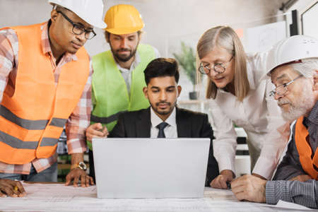 Four men and one woman sitting together at table and discussing blueprints on laptop during business conference. Multi ethnic architects, designers and engineers working at office.の写真素材