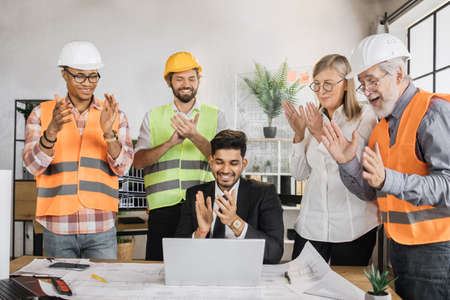 Team of multinational engineers rejoices clapping their hands looking at laptop after a successful project. Hindu boss sitting at table while his colleague engineers are standing around him.の写真素材