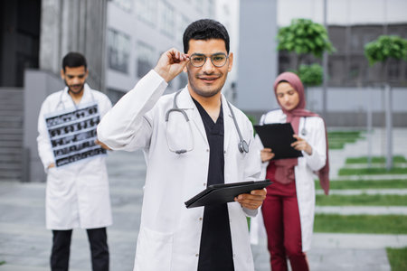 Coworking outdoors of hospital. Young handsome arab hindu man doctor in glasses using tablet device, posing to camera with smile. Multi ethnic male and female physicians working on the backgroundの写真素材