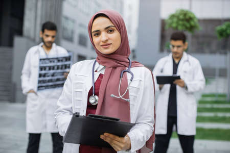 Close up of female Muslim pleasant doctor in hijab, standing with clipboard outdoors in front of hospital building, while two handsome Arabian men doctors working with x-ray and tablet pc behindの写真素材