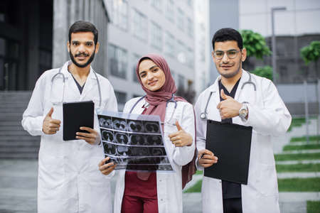 Portrait of three young Arabian doctors, posing to camera outdoors in front of modern clinic building, showing thumbs up. Radiologist doctors holding x-ray, clipboard and tablet pc.の写真素材