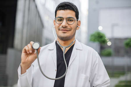 Portrait of smiling friendly male Asian doctor or medical student in eyeglasses, standing outside modern hospital, demonstrating his stethoscope to camera. Focus on faceの写真素材