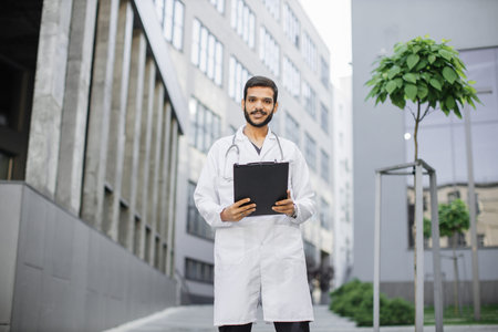 Portrait of young handsome bearded Indian man physician healthcare worker in white coat, standing...